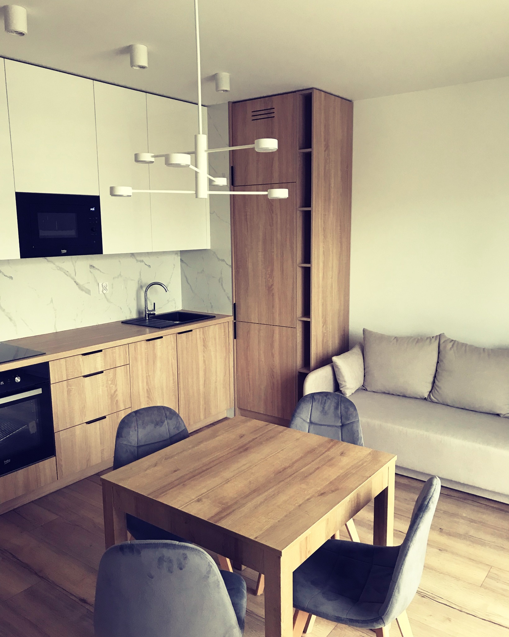 Nowoczesne wnętrze: drewniany stół z szarymi krzesłami na tle jasnej kuchni z marmurowym backsplashiem i drewnianymi szafkami oraz beżowej sofy.