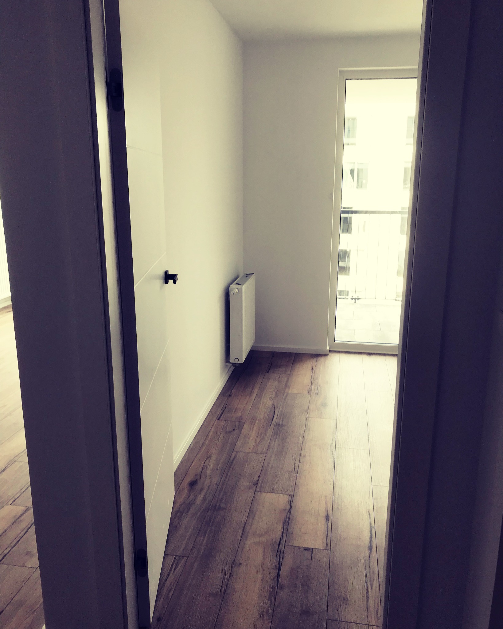 Widok zza uchylonych drzwi na pokój z drewnopodobną podłogą, białymi ścianami, grzejnikiem i balkonem z widokiem na budynek.