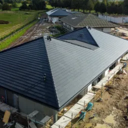 Nowoczesny dom z ciemnoszarą dachówką ceramiczną, widok z góry na świeżo położony dach, rusztowania wokół budynku, w tle pola i inne budynki w trakcie budowy.