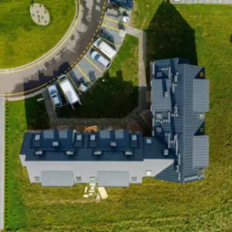 Widok z drona na budynek z ciemnoszarym dachem, obok zaparkowane samochody i materiały budowlane na trawie, sugerujące prace dekarskie.