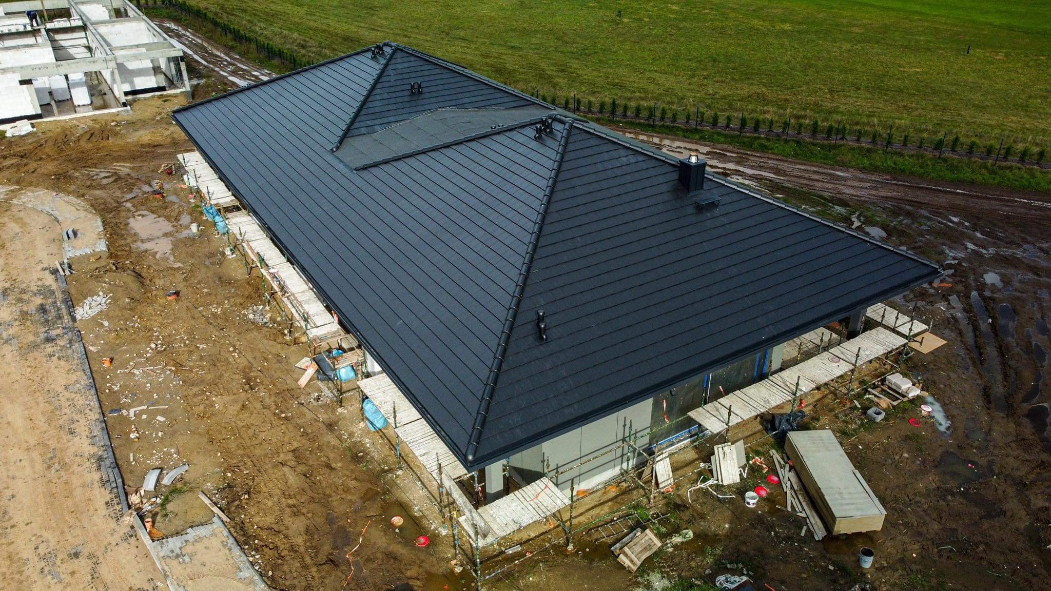 Nowoczesny dom w trakcie budowy z ciemnoszarym, płaskim dachem, widok z góry na rusztowania i teren budowy.