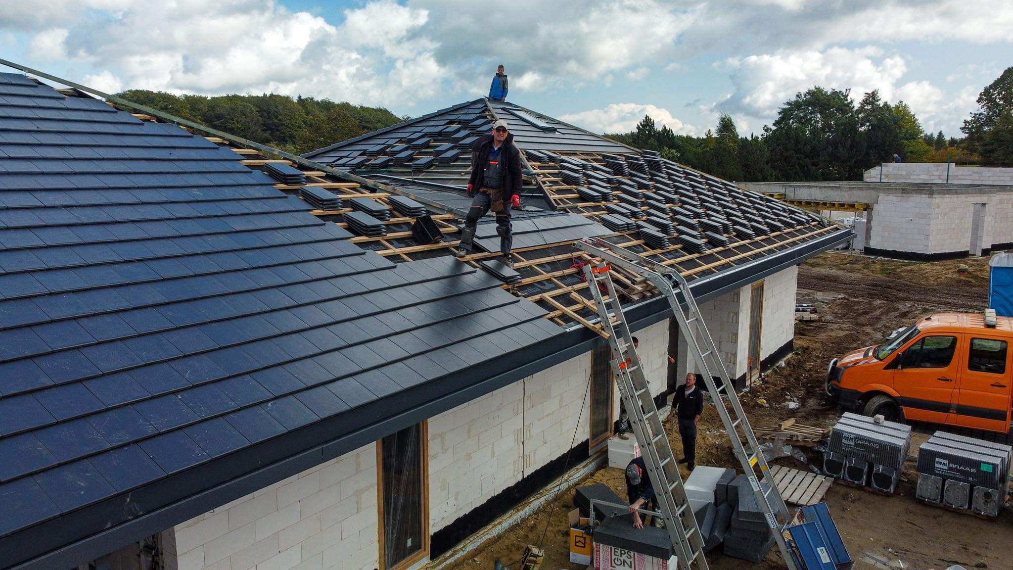 Montaż czarnej dachówki ceramicznej na nowym domu jednorodzinnym; dekarz w okularach słonecznych stoi na dachu, a drugi pracuje przy drabinie, widoczne palety z dachówką BMI Braas i pomarańczowy...