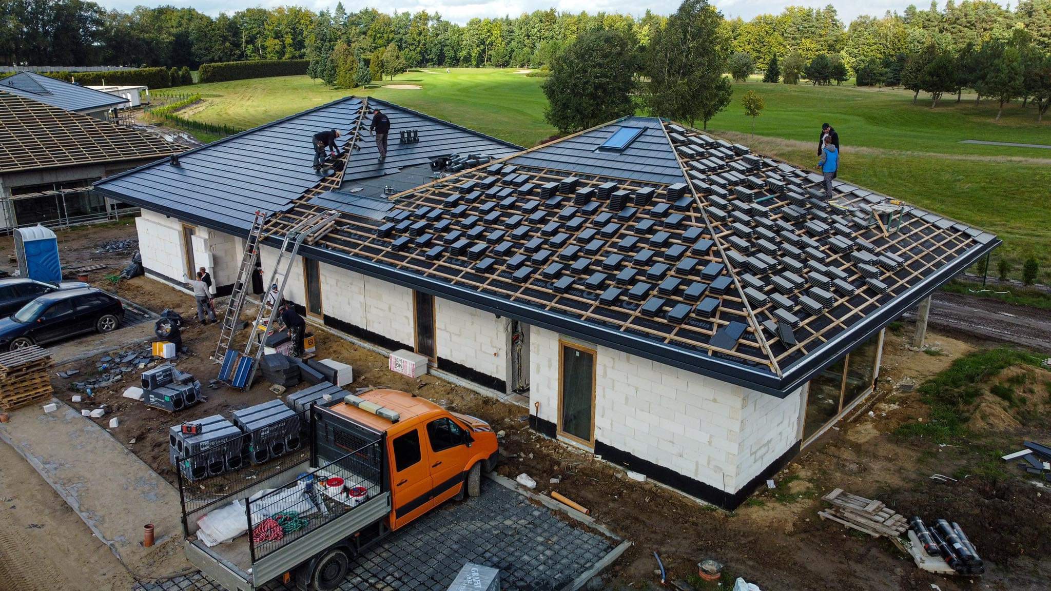 Montaż czarnej dachówki na nowym domu z białego bloczka, widok z góry na częściowo pokryty dach, pomarańczowy samochód dostawczy i pracownicy na dachu.