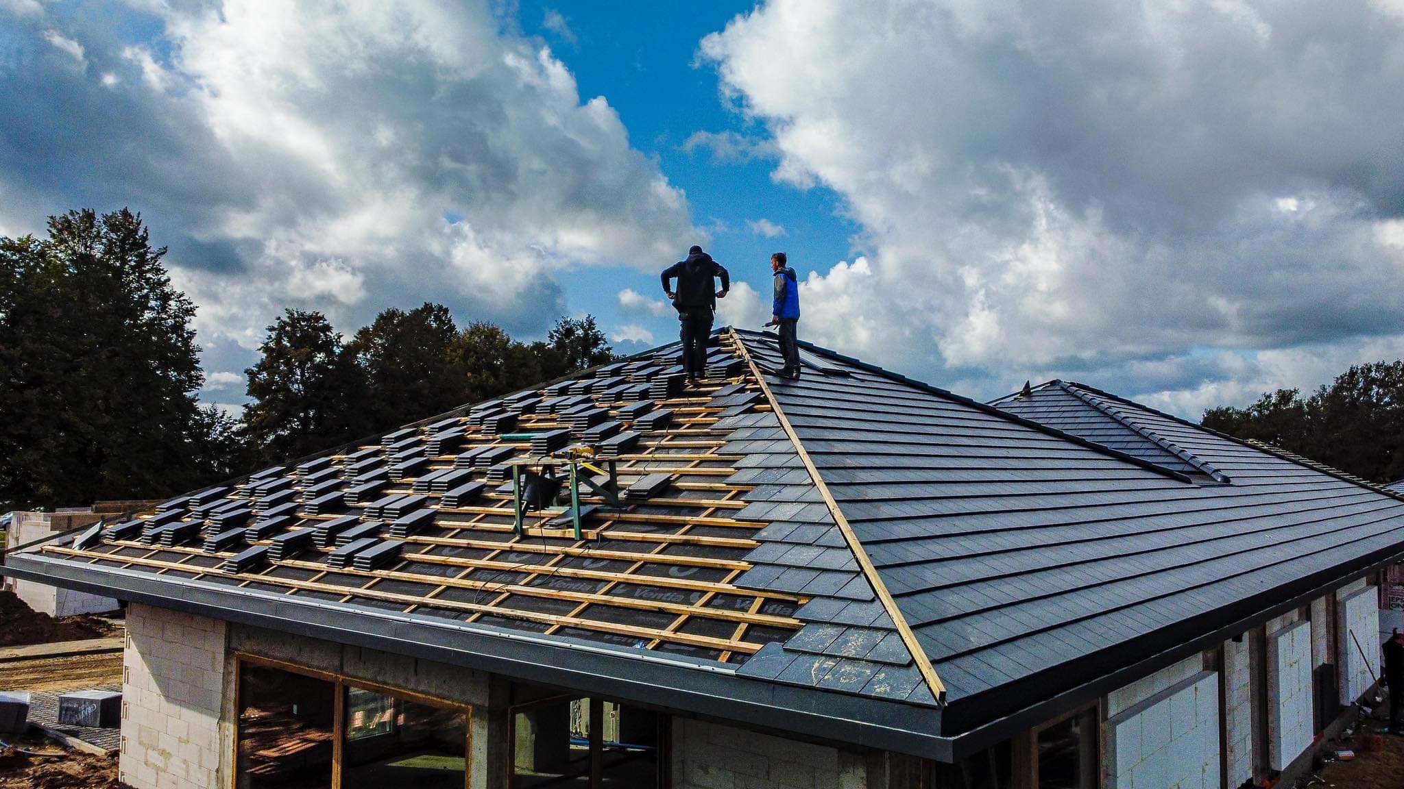 Dwóch dekarzy pracuje na szczycie dachu domu jednorodzinnego, układając ciemnoszarą dachówkę ceramiczną na drewnianej konstrukcji, widok z góry na tle pochmurnego nieba.