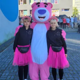 Dwie kobiety w czarnych bluzach i różowych tiulowych spódnicach, z kwiatowymi wiankami na głowach, pozują obok dużej maskotki różowego misia na tle szarego budynku.