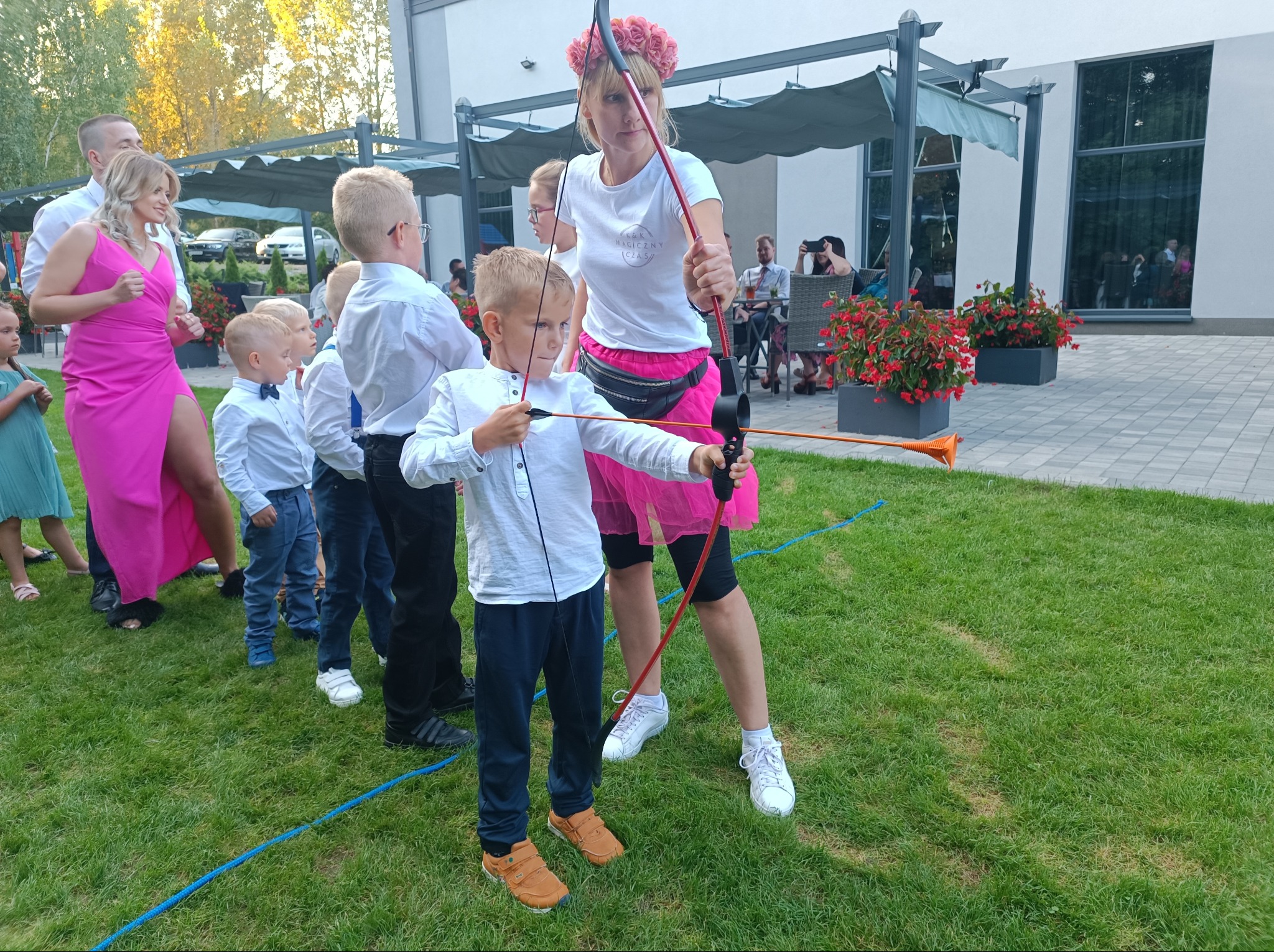 Grupa dzieci ustawiona w kolejce, obserwuje jak chłopiec celuje z łuku pod okiem kobiety w różowej spódnicy i wianku na głowie, na trawniku przed budynkiem.