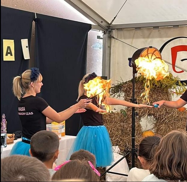 Kobiety w niebieskich tiulowych spódnicach prezentują pokaz z ogniem na tle dekoracji ze słomy i dyni, widownia obserwuje.