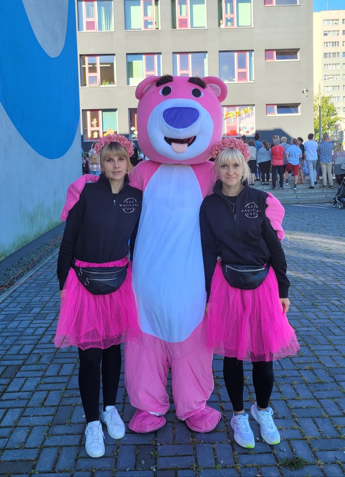 Dwie kobiety w czarnych bluzach i różowych tiulowych spódnicach, z kwiatowymi wiankami na głowach, pozują obok dużej maskotki różowego misia na tle szarego budynku.