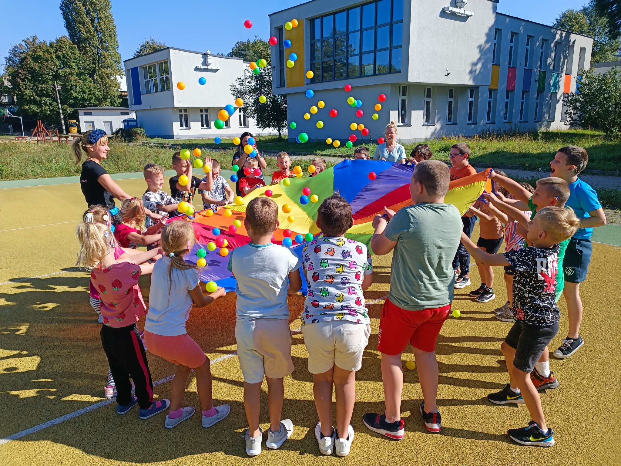 Grupa dzieci trzymająca kolorową płachtę, podrzucająca w górę kolorowe piłeczki na tle budynku szkolnego i drzew.