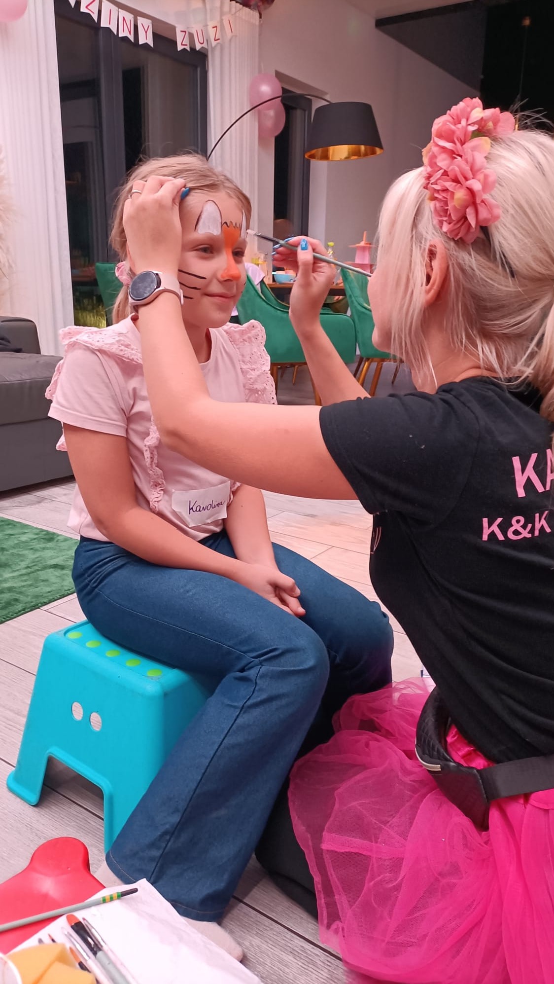 Dziewczynka siedzi na turkusowym stołku, podczas gdy artystka maluje jej twarz w motyw lisa, używając cienkiego pędzla; widoczny napis 'Karolina' na bluzce dziewczynki, a artystka ma kwiaty we...