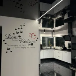 Nowoczesna kuchnia z czarno-białymi meblami, czarną podłogą imitującą marmur i białym sufitem podwieszanym z oświetleniem LED, na ścianie dekoracyjny napis 'Dom tworzy Rodzina' z motywem motyli...