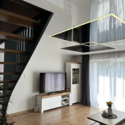 Błyszczący sufit z podświetleniem LED w kształcie kwadratów odbija wnętrze salonu z drewnianymi schodami, telewizorem i oknem z zasłonami.