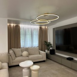 Sufity Napinane Dom Sufitow - Nowoczesny salon z beżową sofą, okrągłym stolikiem i designerską lampą sufitową w kształcie pierścieni. Minimalistyczne wnętrze z telewizorem i roślinami doniczkowymi.