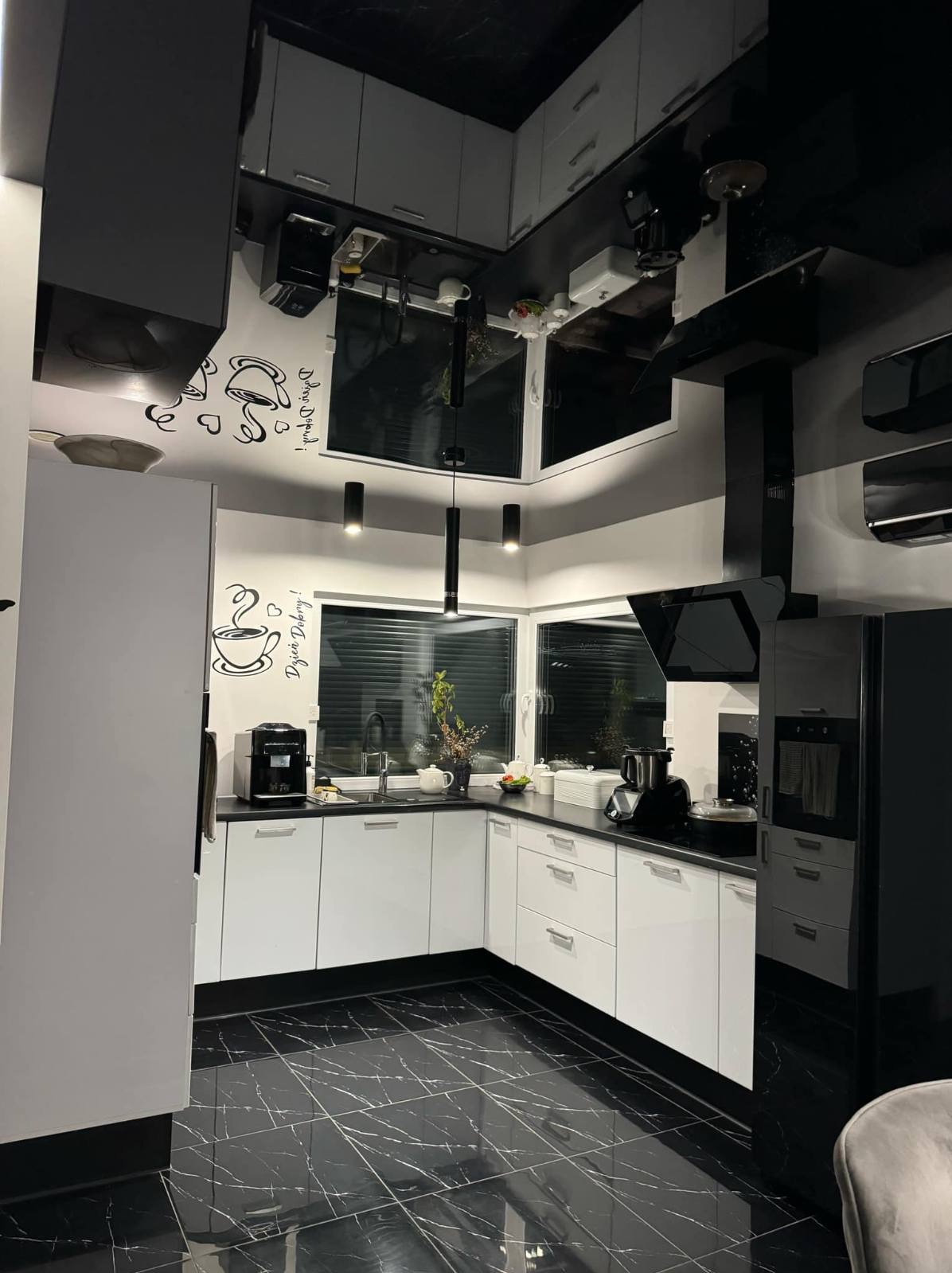 Nowoczesna kuchnia w czerni i bieli z marmurową podłogą, białymi szafkami i czarnymi akcentami, widoczne dekoracyjne naklejki z motywem kawy na ścianie.