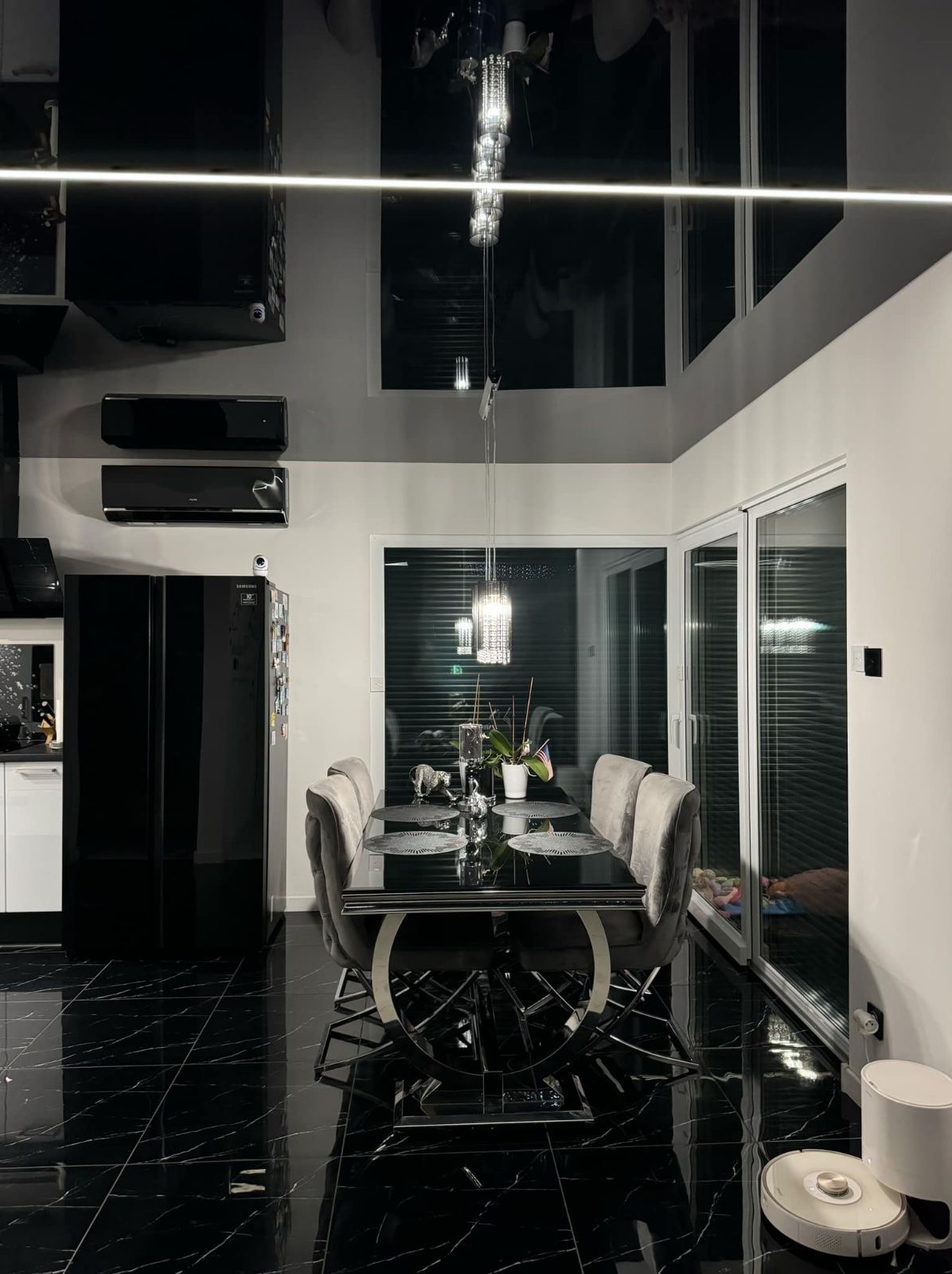 Nowoczesna jadalnia z błyszczącym czarnym sufitem napinanym, odbijającym żyrandol i światło LED, czarna podłoga z białymi żyłami, stół z szarymi krzesłami i czarny sprzęt AGD.