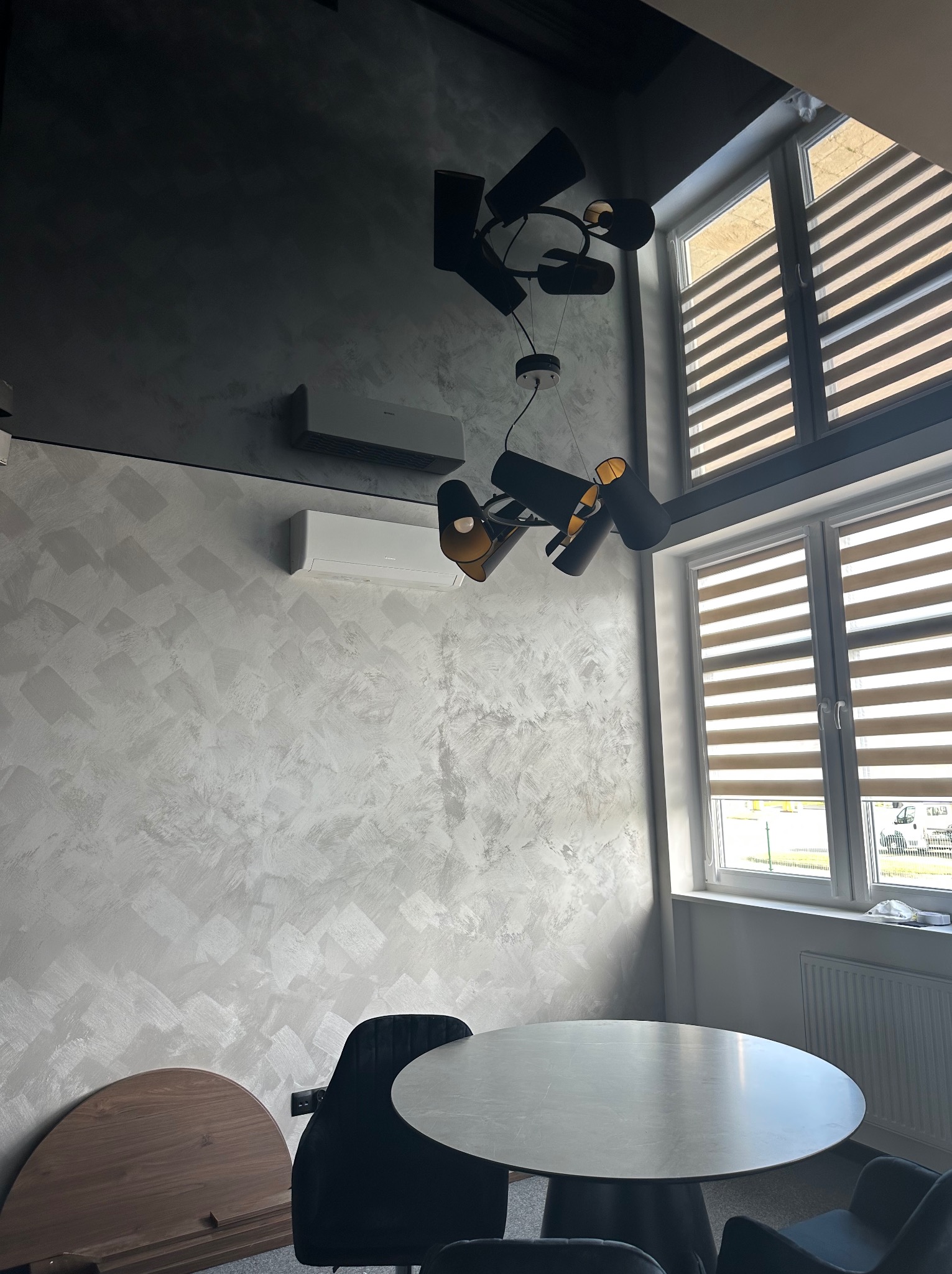 Nowoczesne wnętrze z szarą, teksturowaną ścianą, designerską lampą wiszącą, dwoma klimatyzatorami i oknami z żaluzjami poziomymi. Widoczny okrągły stół i krzesła.