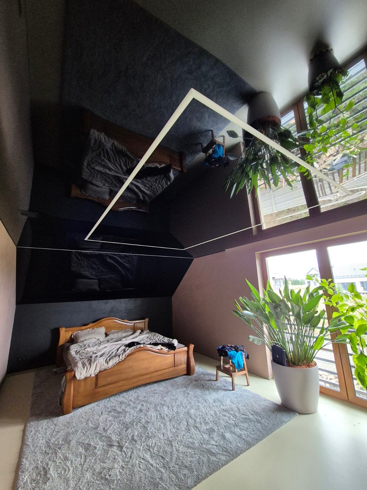 Sypialnia na poddaszu z drewnianym łóżkiem i szarą wykładziną, czarny błyszczący sufit z oświetleniem LED odbija wnętrze, widoczne rośliny doniczkowe przy oknie.