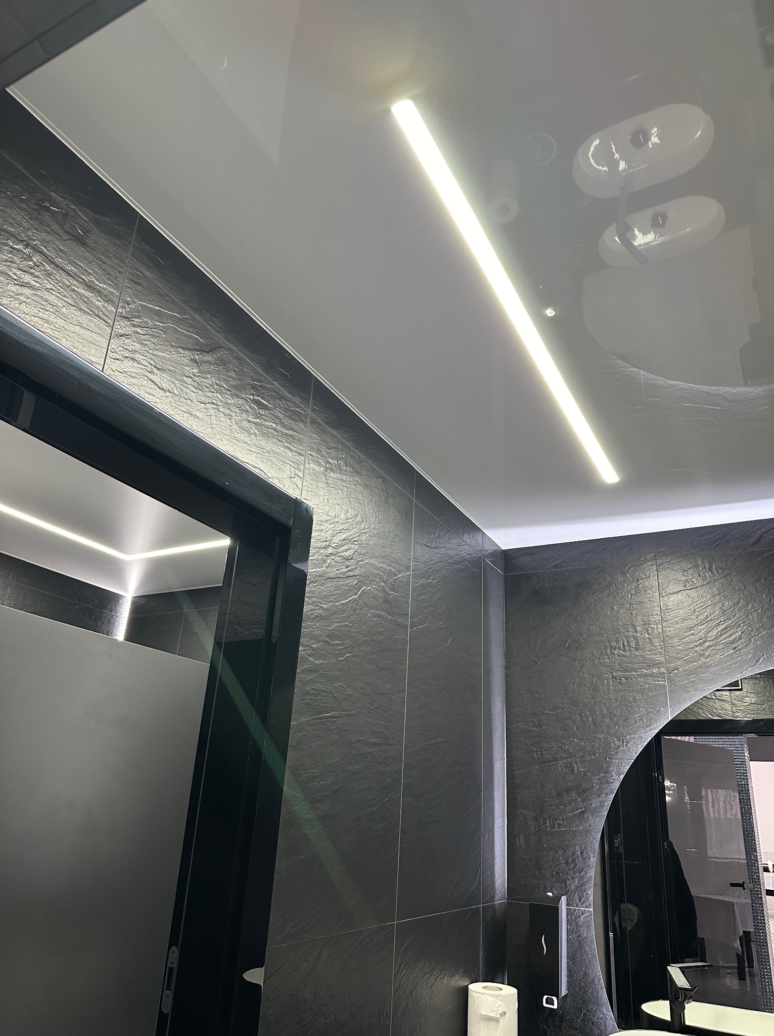 Biały, błyszczący sufit napinany z liniowym oświetleniem LED w nowoczesnej łazience z czarnymi płytkami imitującymi kamień i lustrem o zaokrąglonym kształcie.
