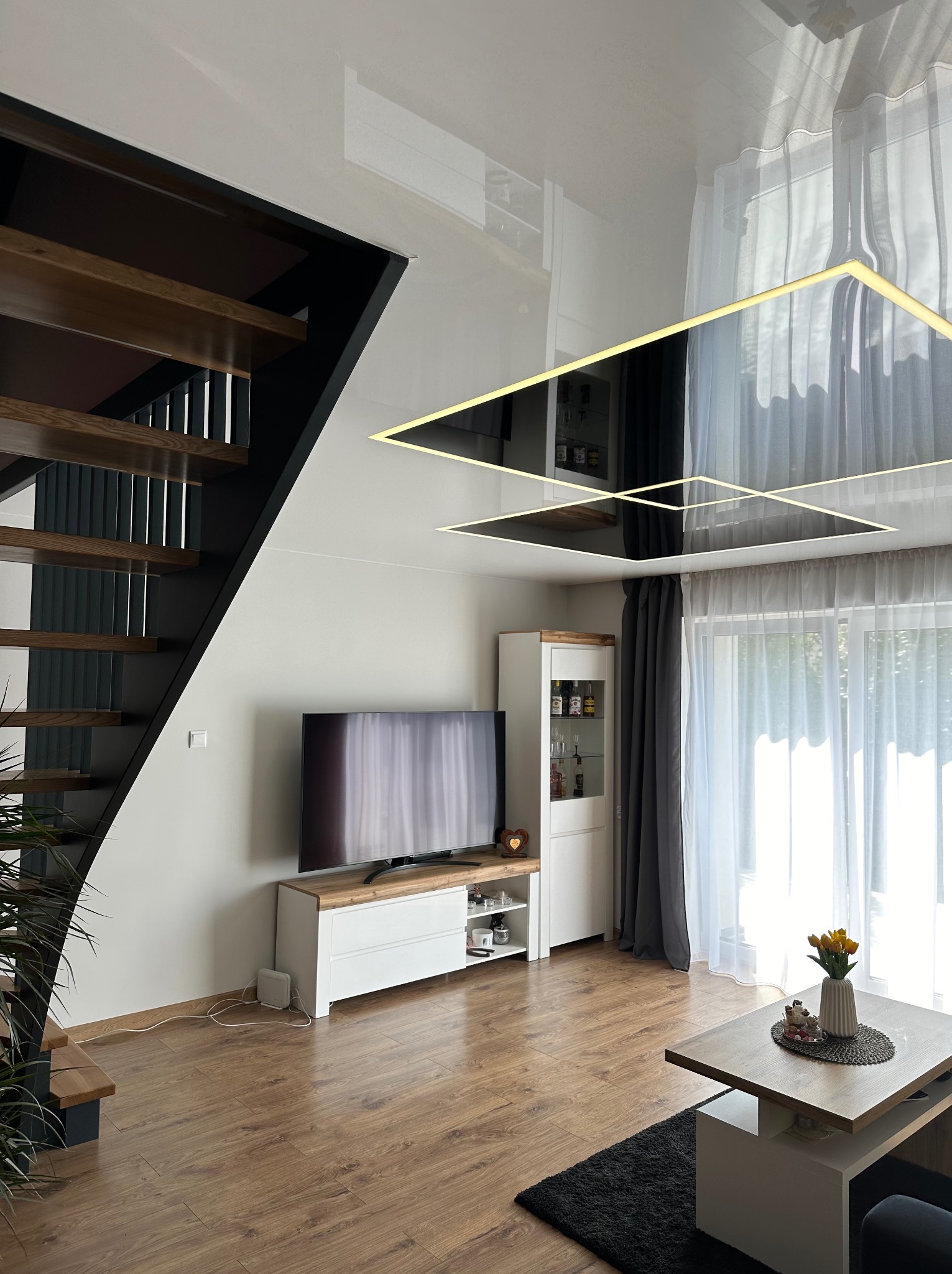 Błyszczący sufit z podświetleniem LED w kształcie kwadratów odbija wnętrze salonu z drewnianymi schodami, telewizorem i oknem z zasłonami.