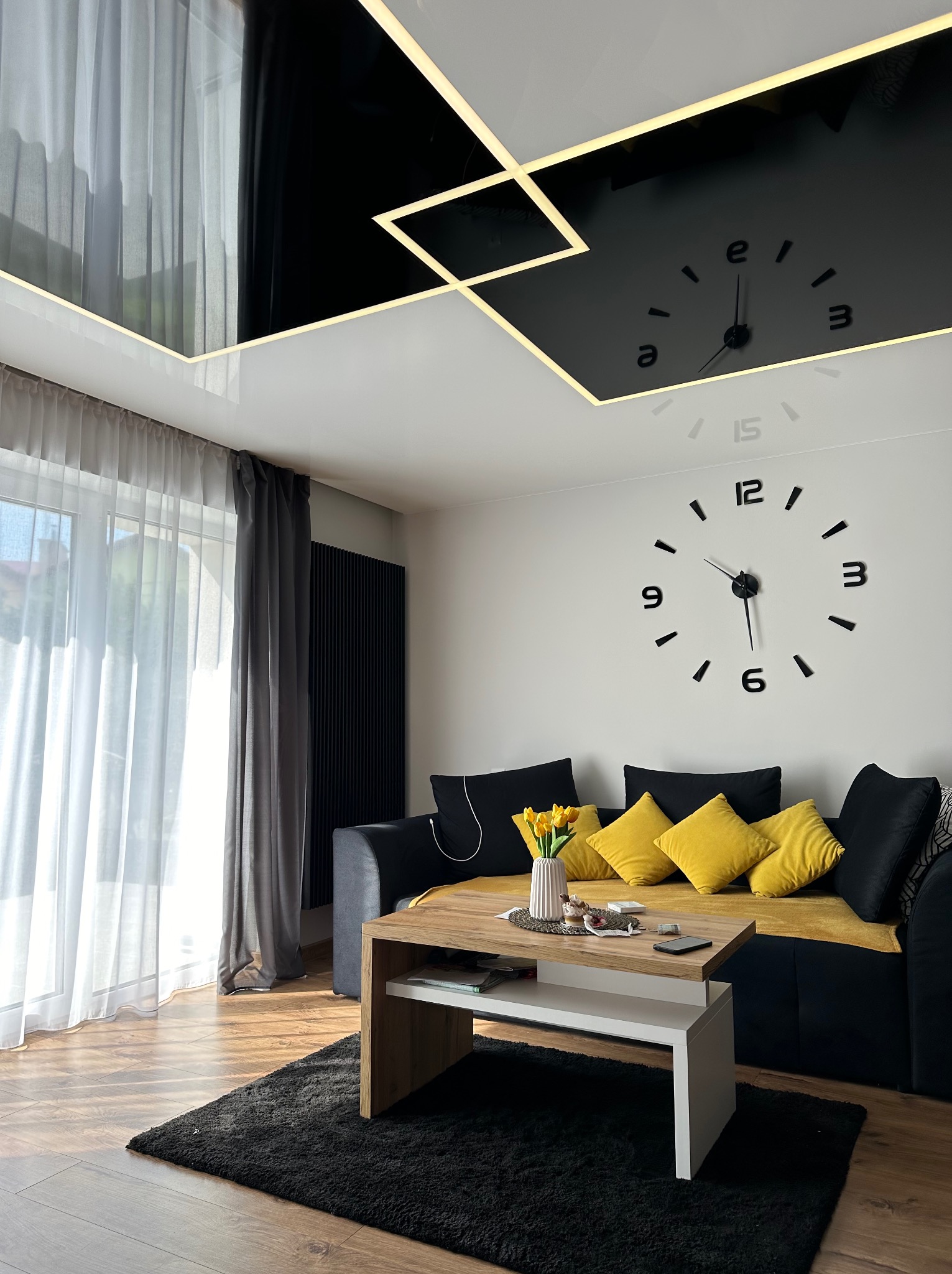 Nowoczesny salon z czarnym, błyszczącym sufitem z geometrycznym oświetleniem LED i dużym zegarem ściennym, żółte poduszki na czarnej sofie, bukiet tulipanów na stoliku kawowym.
