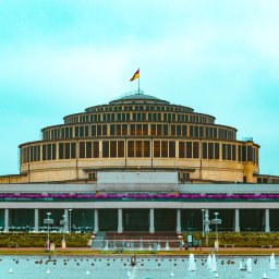 Wojciech Kosela Photography - Hala Stulecia we Wrocławiu sfotografowana w Pile. Widoczne fontanny, budynek z flagą na szczycie i transparenty z informacjami turystycznymi.