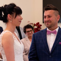 Wojciech Kosela Photography - Para młoda wymienia spojrzenia podczas ceremonii ślubnej, panna młoda w koronkowej sukni, pan młody w granatowym garniturze w kratę z różową muszką, w tle uśmiechnięta kobieta.