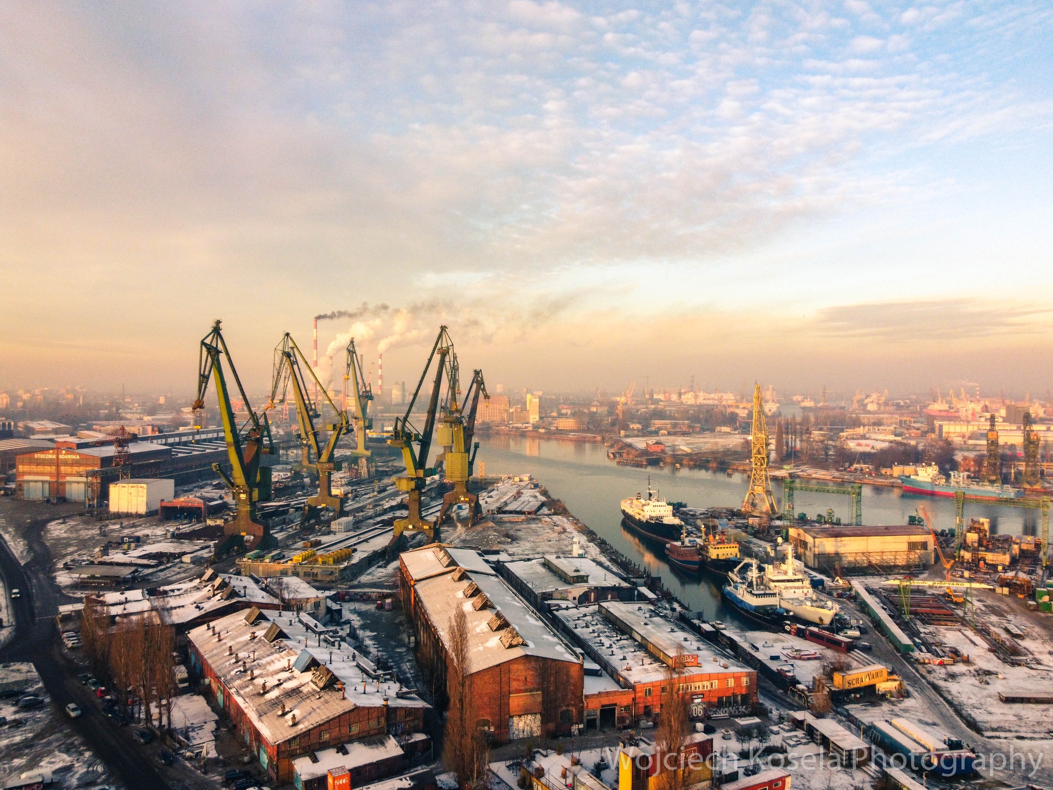 Panorama stoczni w Gdańsku z lotu ptaka, widoczne dźwigi portowe, budynki pokryte śniegiem i statki w dokach, w tle kominy i zabudowa miejska w porannym świetle.