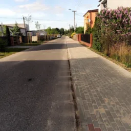 Ulica z chodnikiem wyłożonym szarą kostką brukową, obrzeża z czerwonej kostki, po drugiej stronie jezdni asfaltowa droga, w tle domy i drzewa.