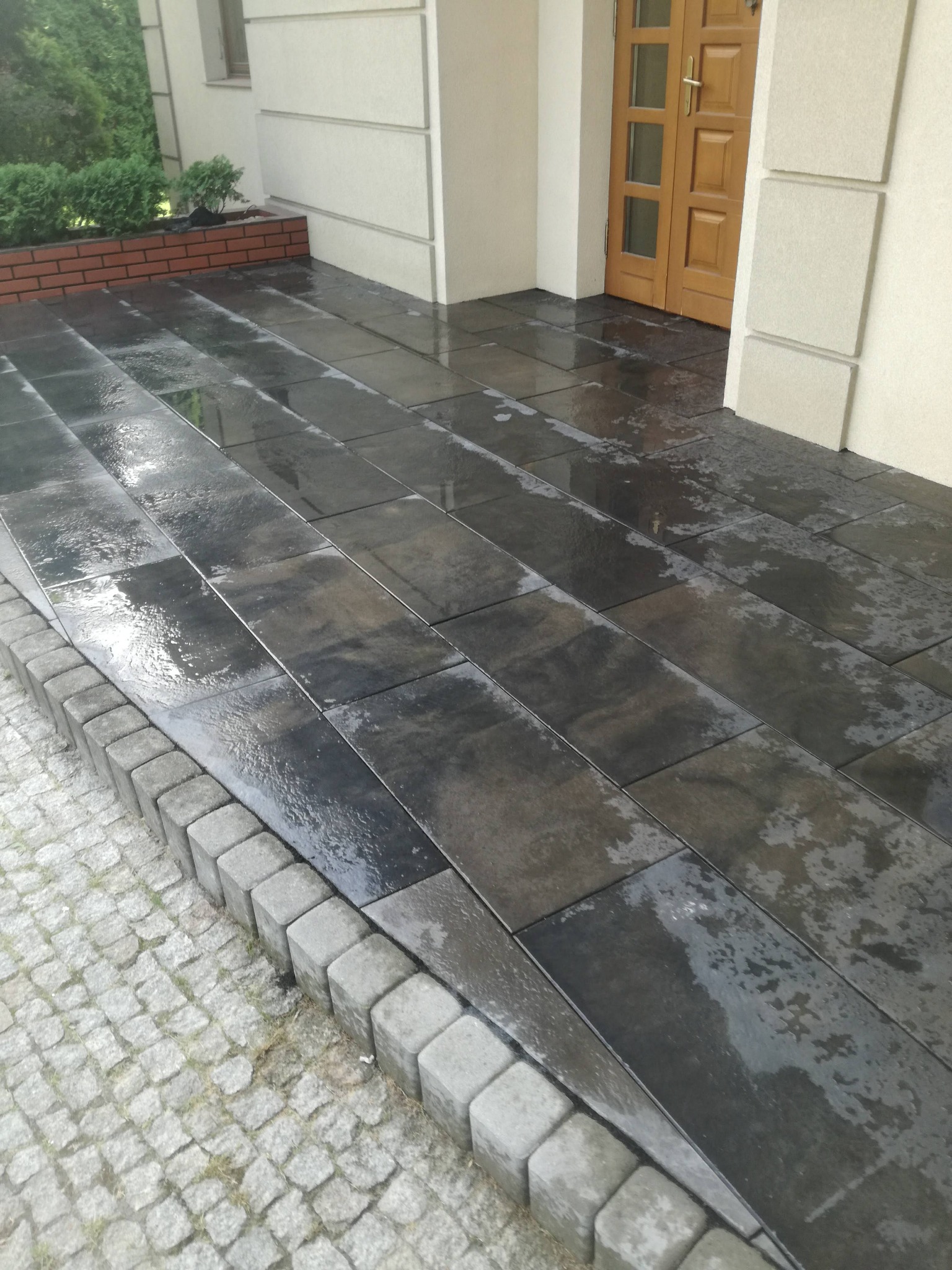 Świeżo ułożona, mokra, ciemnoszara nawierzchnia z prostokątnych płyt na podjeździe, obramowana kostką brukową, prowadząca do drzwi wejściowych budynku.