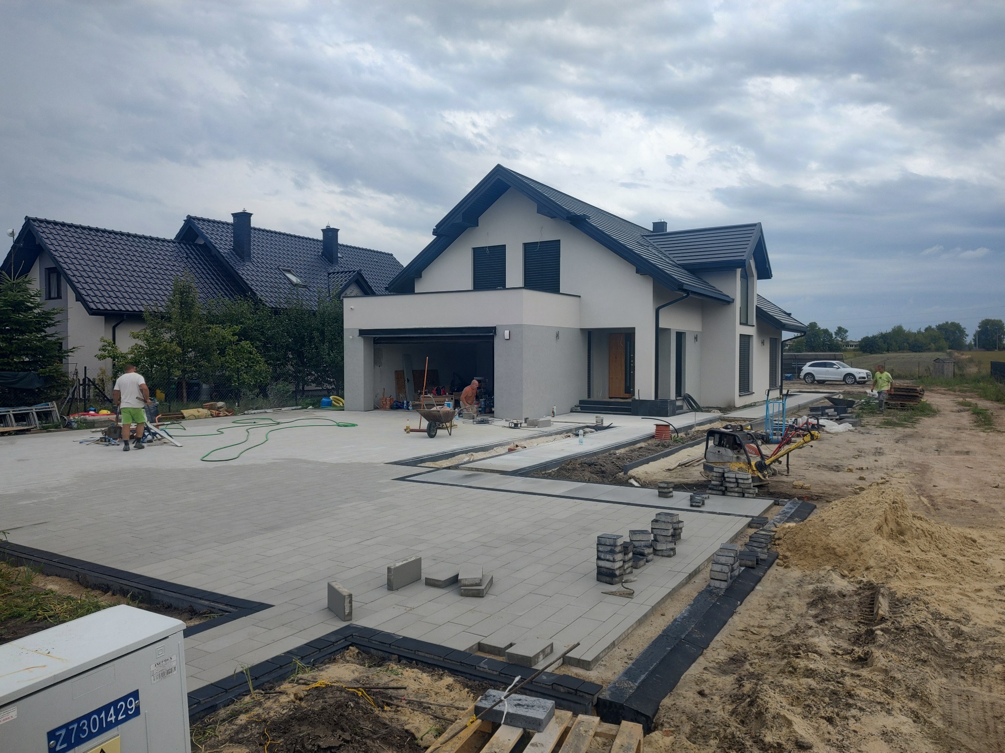 Układanie szarej kostki brukowej na podjeździe do nowoczesnego domu z garażem, widoczne narzędzia i materiały budowlane, praca w toku.