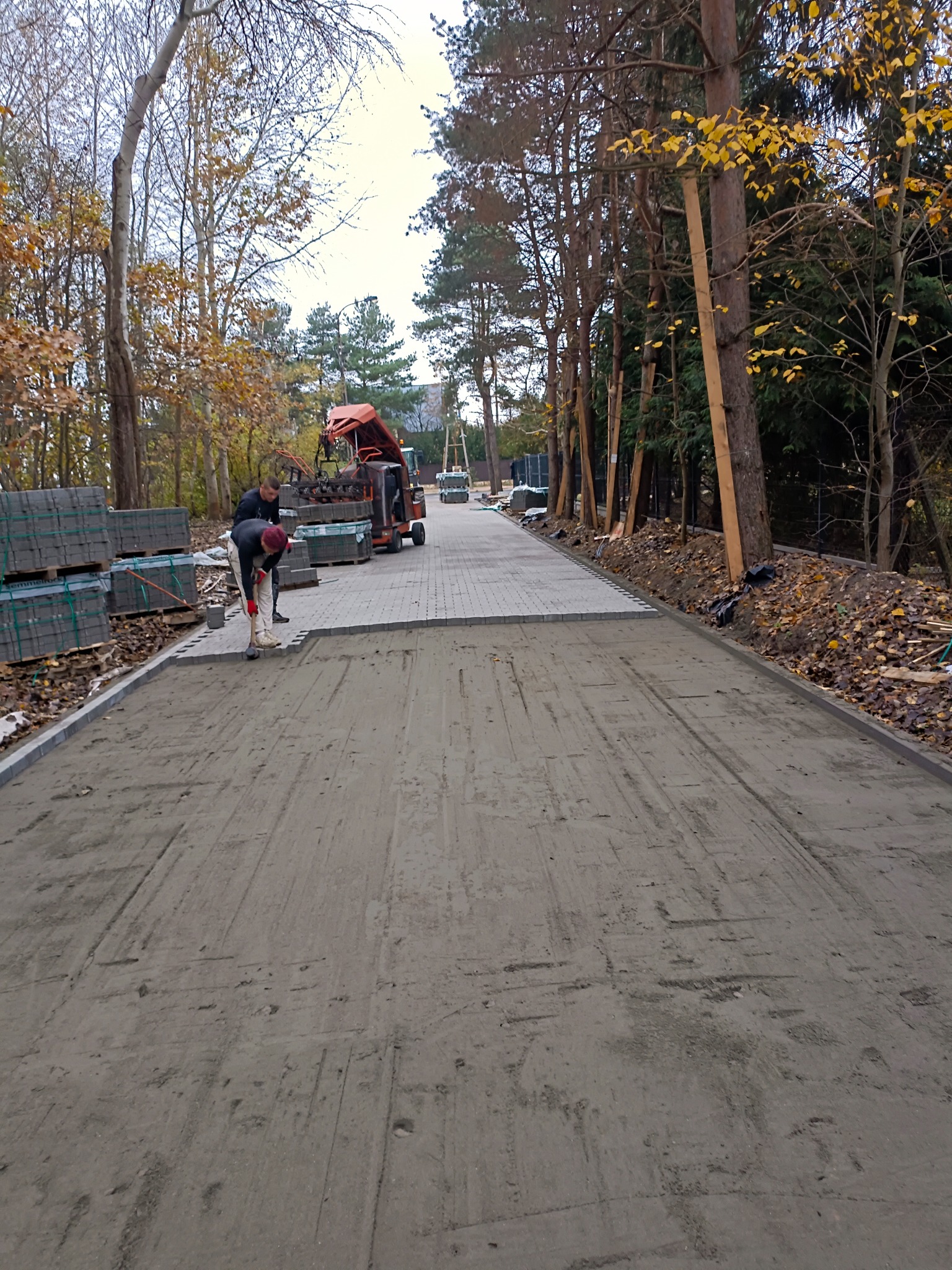 Pracownik ubija kostkę brukową na przygotowanym podłożu piaskowym, w tle palety z kostką i maszyna do układania kostki, teren częściowo zadrzewiony.
