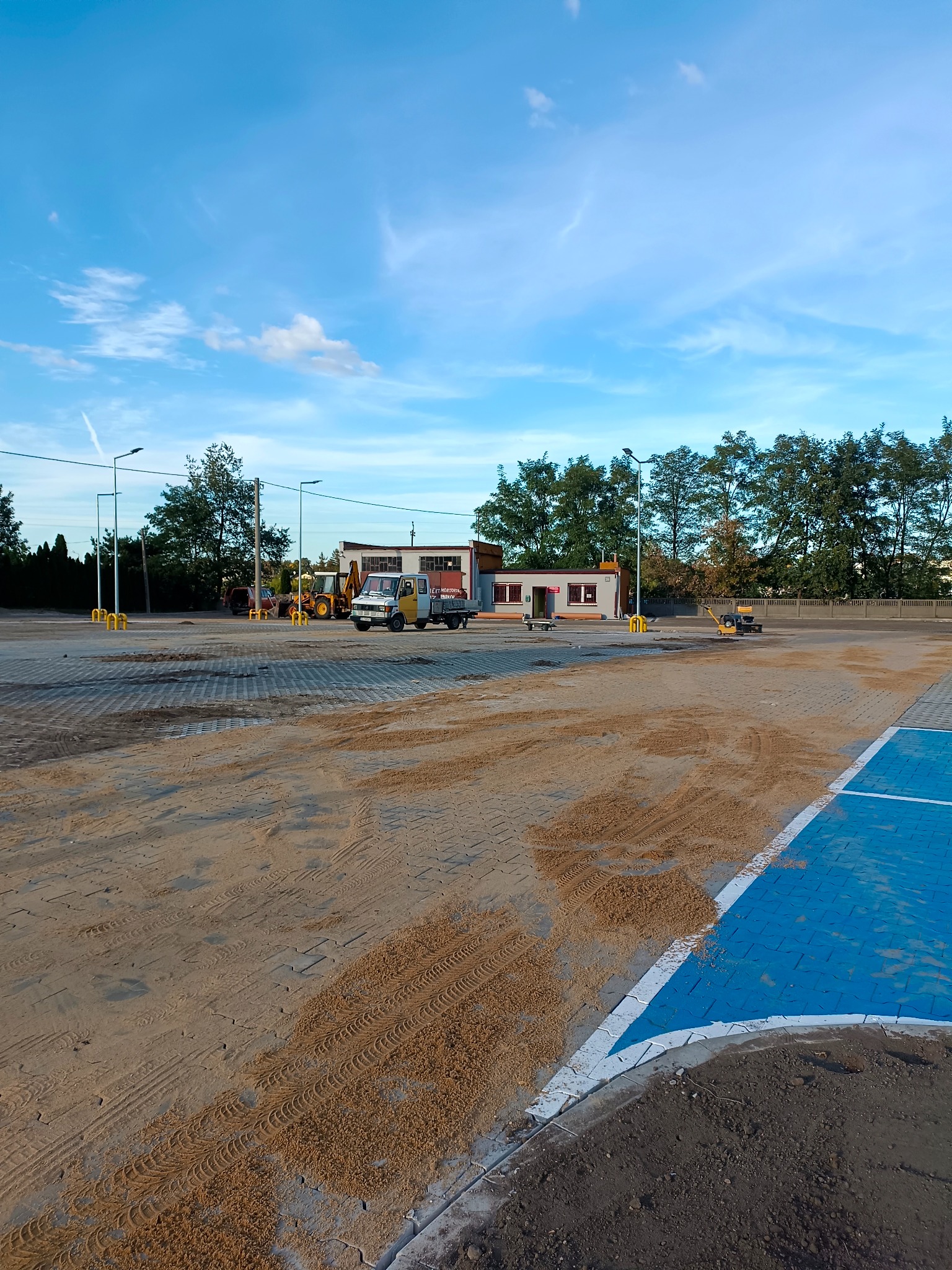 Rozległy plac z częściowo ułożoną kostką brukową, pokrytą piaskiem, z widocznymi śladami opon, w tle budynek, drzewa i maszyny budowlane, w tym żółta koparka i biało-żółty samochód dostawczy.