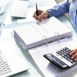 MarWika Biuro Rachunkowe Maria Jóskowska - Kobieta przy biurku, kalkulator, laptop, dokumenty w segregatorze, monitor. Ręka pisząca długopisem, druga obsługująca kalkulator. Jasne biuro, praca biurowa.