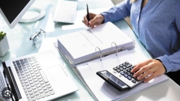 Kobieta przy biurku, kalkulator, laptop, dokumenty w segregatorze, monitor. Ręka pisząca długopisem, druga obsługująca kalkulator. Jasne biuro, praca biurowa.