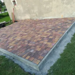 Ułożona kostka brukowa w odcieniach brązu i beżu na przygotowanym podłożu, obrzeża betonowe, widoczny fragment trawnika i ściana budynku w tle, wózek budowlany z lewej strony.