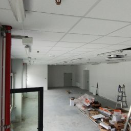 Łukasz Gabrys - Wnętrze w trakcie prac wykończeniowych, widoczne drabiny, materiały budowlane i sufit kasetonowy z brakującymi panelami.