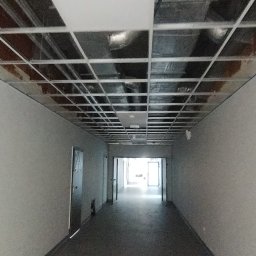 Łukasz Gabrys - Korytarz w trakcie prac wykończeniowych, widoczna metalowa konstrukcja sufitu podwieszanego i świeżo pomalowane ściany na szaro.