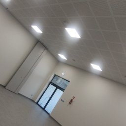 Łukasz Gabrys - Nowoczesne wnętrze z gładkimi, jasnymi ścianami, podwieszanym sufitem z oświetleniem LED i szarą podłogą, widok na drzwi wejściowe.