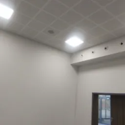 Jasne wnętrze z kasetonowym sufitem podwieszanym, kratką wentylacyjną i widocznym fragmentem ściany z gaśnicą oraz drzwiami.