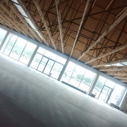 Łukasz Gabrys - Duże pomieszczenie w trakcie prac wykończeniowych: świeżo położone gładzie na ścianach, widoczne rusztowanie z pracownikiem przy suficie, drewniana konstrukcja dachu i duże okna.