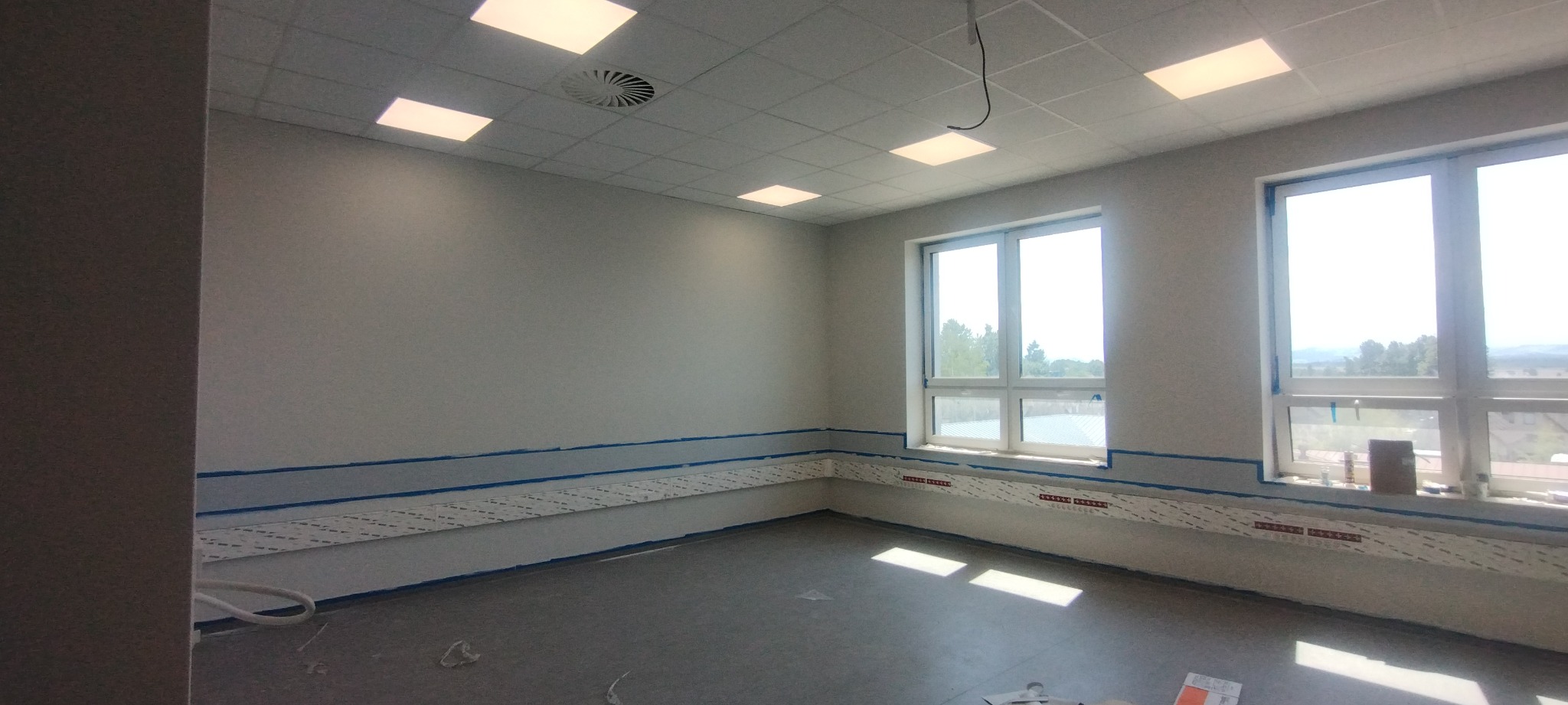 Pokój w trakcie przygotowania do malowania: zabezpieczone ściany taśmą malarską i folią ochronną, sufit z panelami i oświetleniem, widok na zewnątrz przez okna.