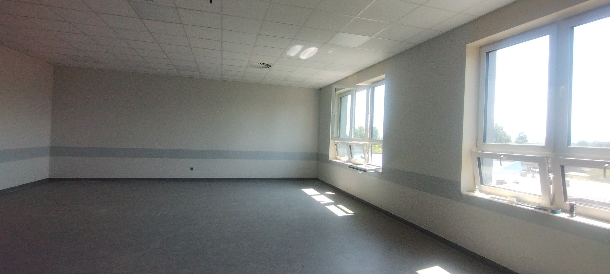 Puste, jasne pomieszczenie z szarą podłogą i białymi ścianami, z widocznymi elementami wykończenia, takimi jak listwy przypodłogowe i sufit podwieszany z kratownicą, częściowo otwarte okna...