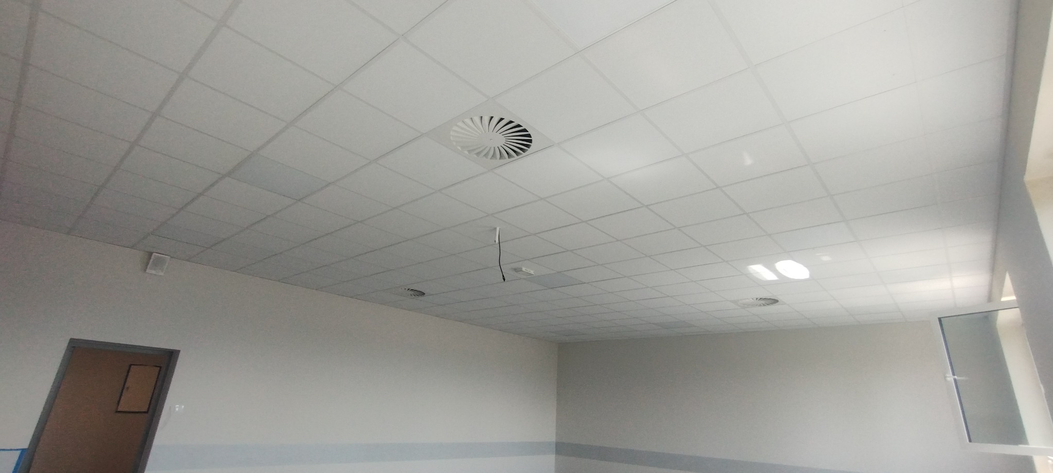Kasetonowy sufit podwieszany z kratką wentylacyjną i wystającym przewodem, widok na fragment pomieszczenia z drzwiami i oknem.