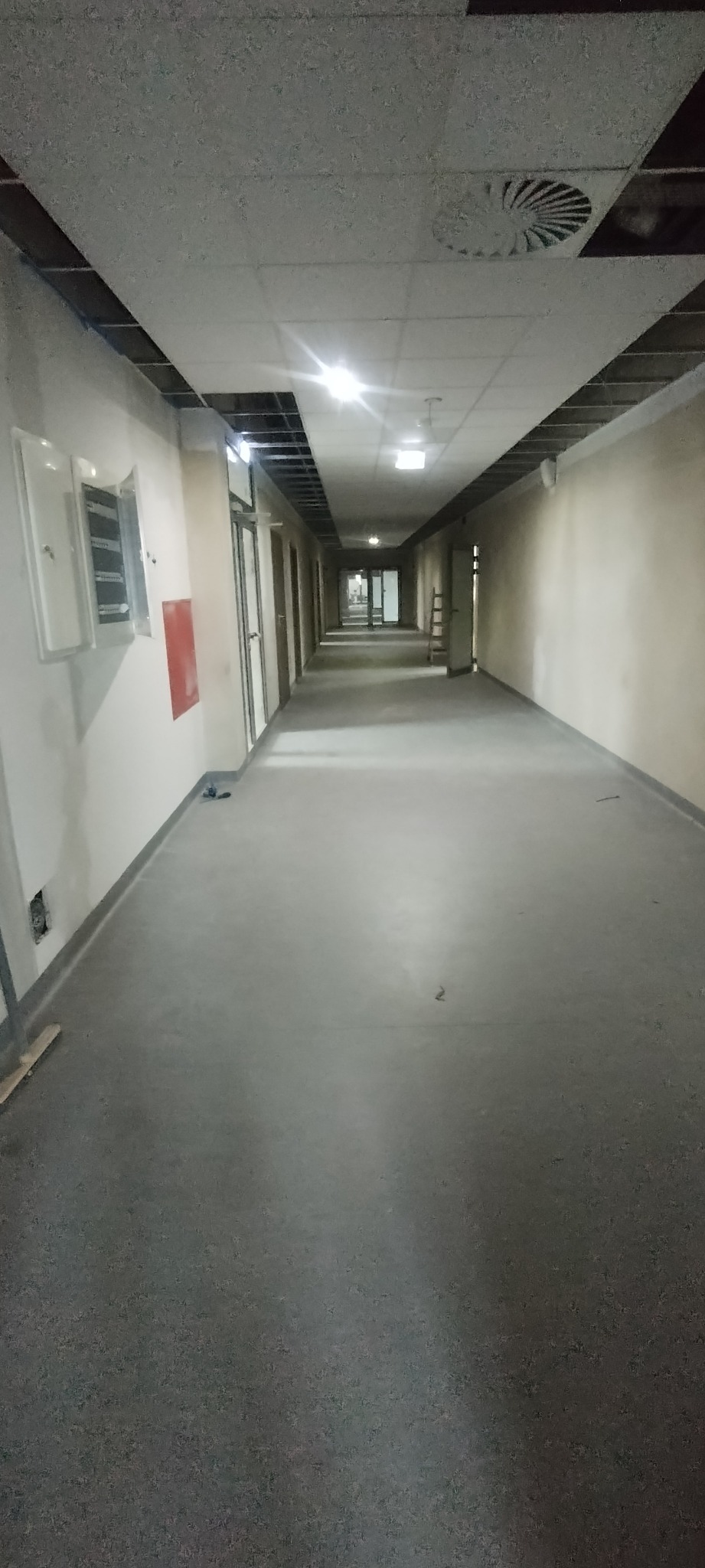 Długi korytarz w trakcie wykańczania, sufit podwieszany z widocznymi elementami konstrukcyjnymi, ściany przygotowane pod malowanie, podłoga z wylewką betonową, widoczne instalacje elektryczne.
