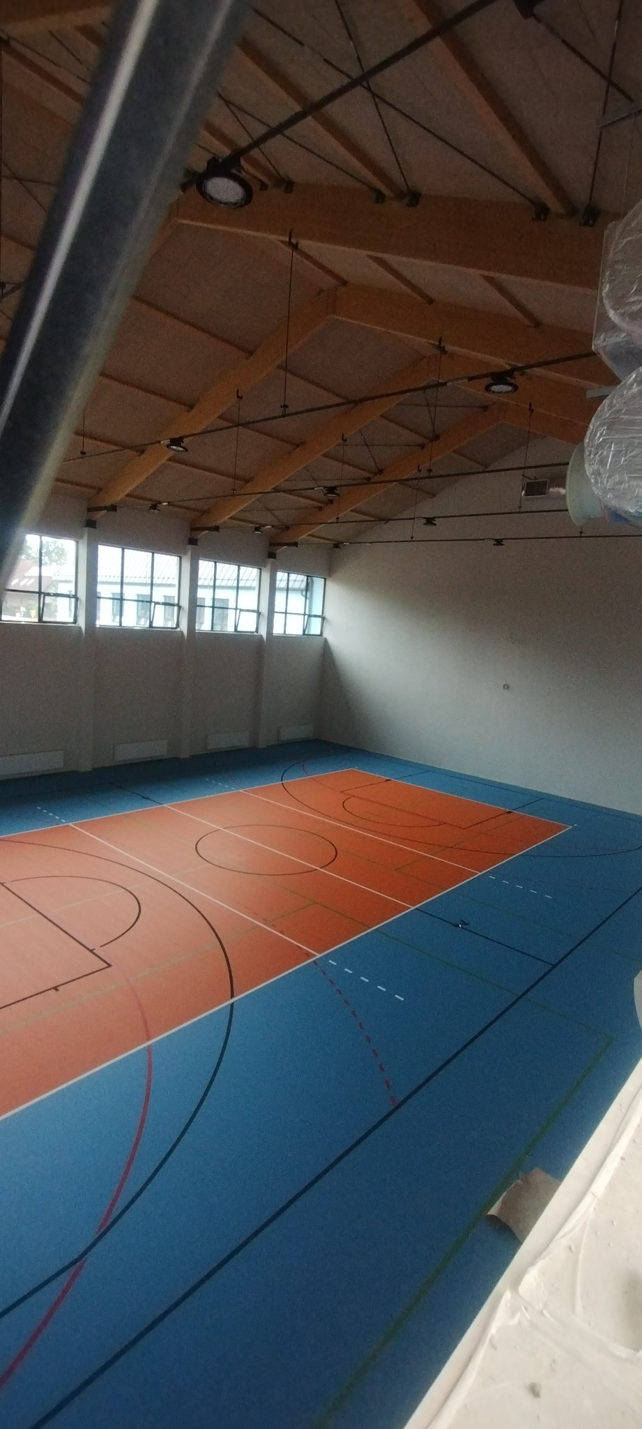 Widok z góry na świeżo pomalowane, puste boisko do koszykówki z niebieską nawierzchnią i pomarańczową strefą gry, oświetlone przez rzędy okien i nowoczesne lampy sufitowe.
