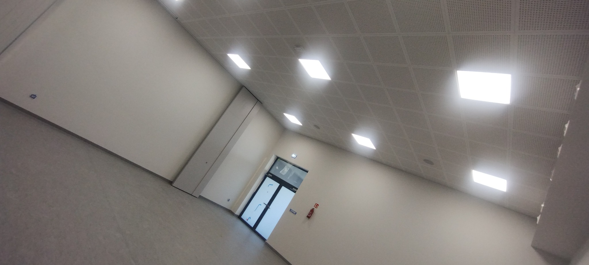 Nowoczesne wnętrze z gładkimi, jasnymi ścianami, podwieszanym sufitem z oświetleniem LED i szarą podłogą, widok na drzwi wejściowe.