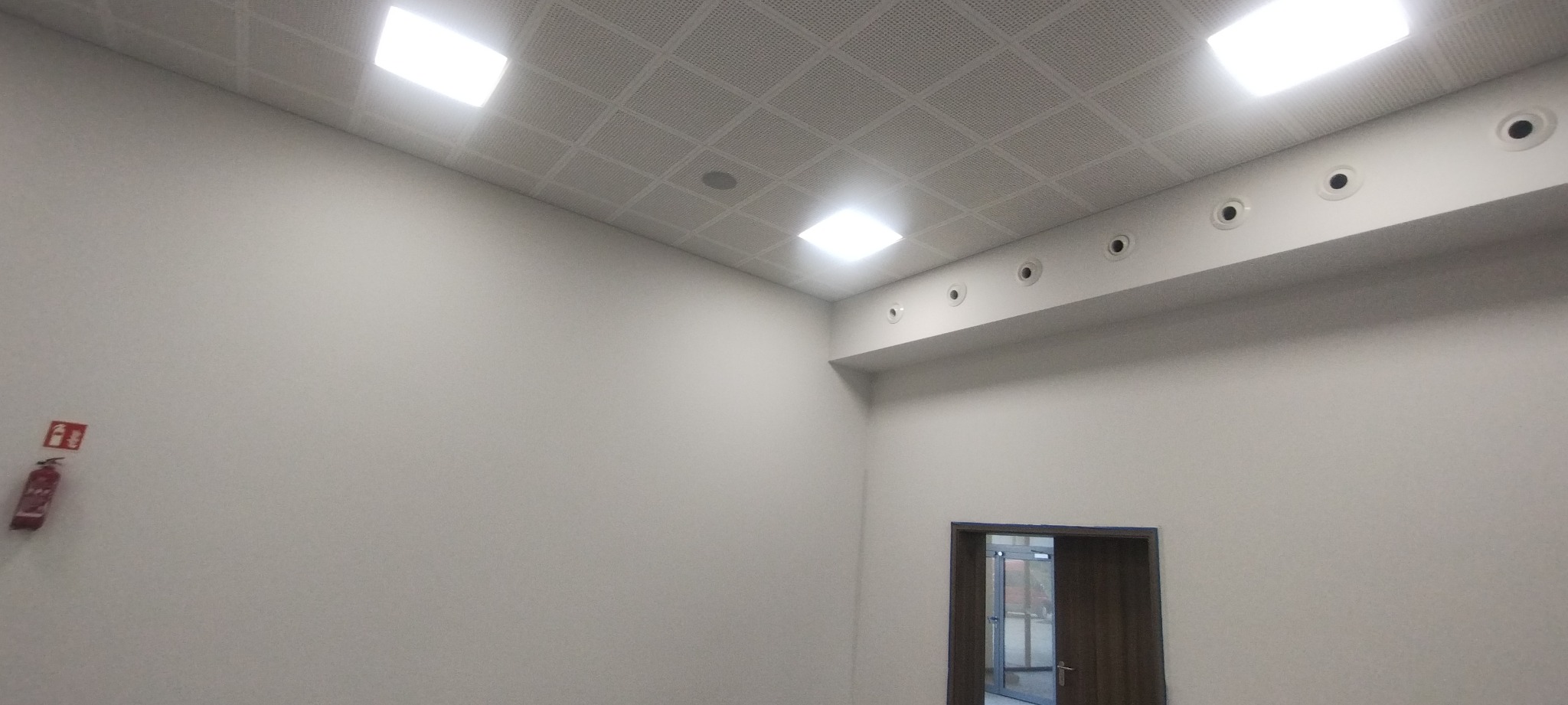 Jasne wnętrze z kasetonowym sufitem podwieszanym, kratką wentylacyjną i widocznym fragmentem ściany z gaśnicą oraz drzwiami.