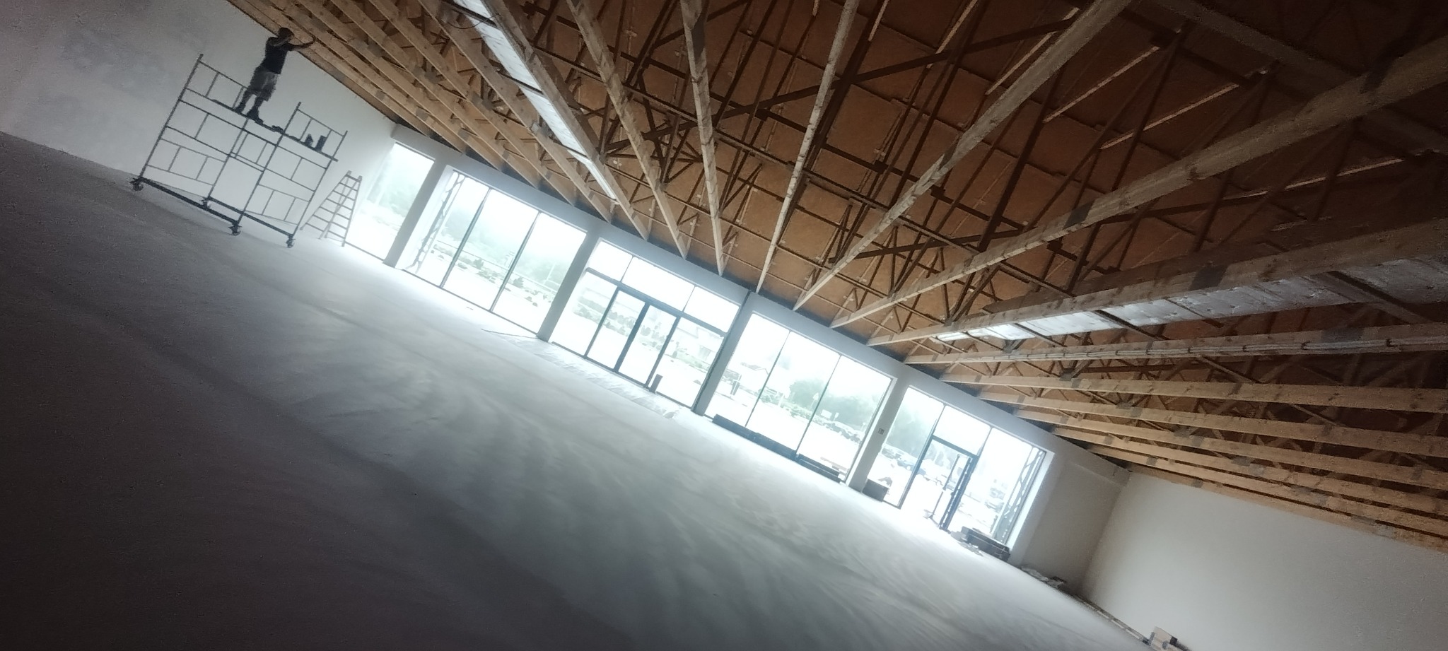 Duże pomieszczenie w trakcie prac wykończeniowych: świeżo położone gładzie na ścianach, widoczne rusztowanie z pracownikiem przy suficie, drewniana konstrukcja dachu i duże okna.