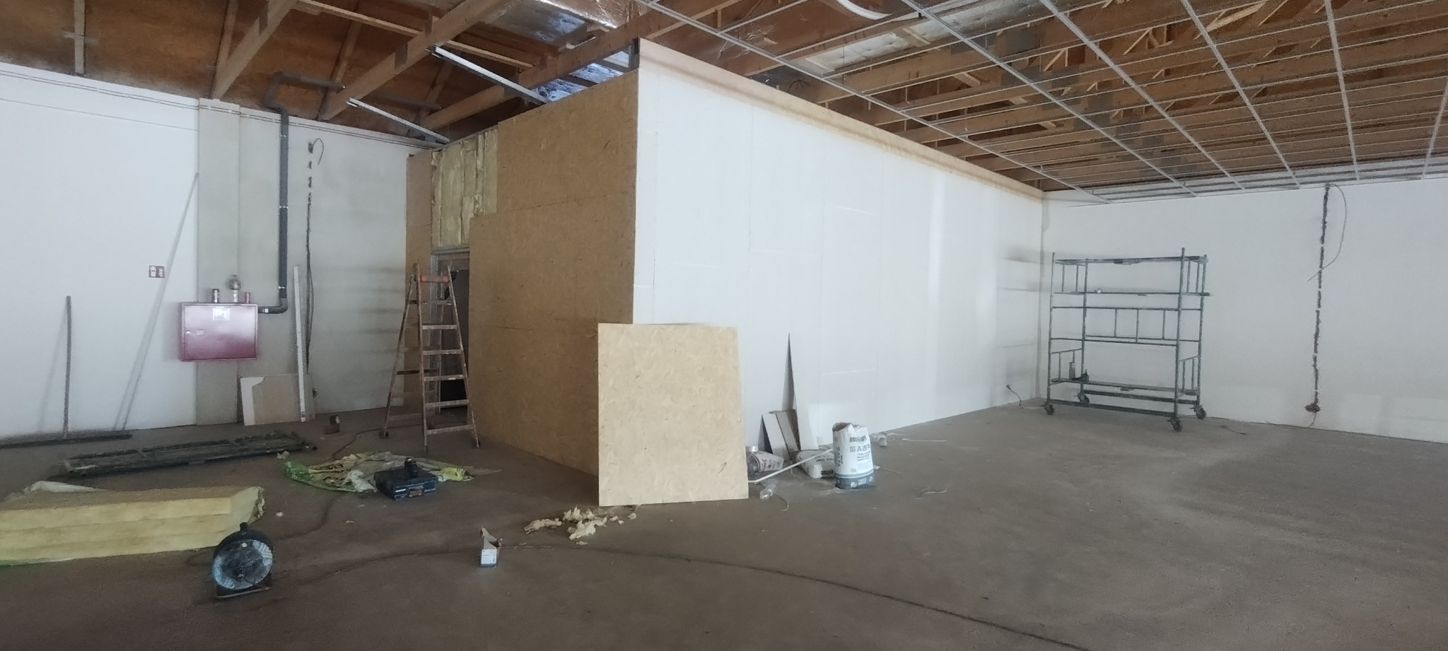 Wnętrze w trakcie adaptacji: ściany z płyt OSB i karton-gipsu, widoczne instalacje pod sufitem, drabina, rusztowanie, materiały izolacyjne i wentylator podłogowy.