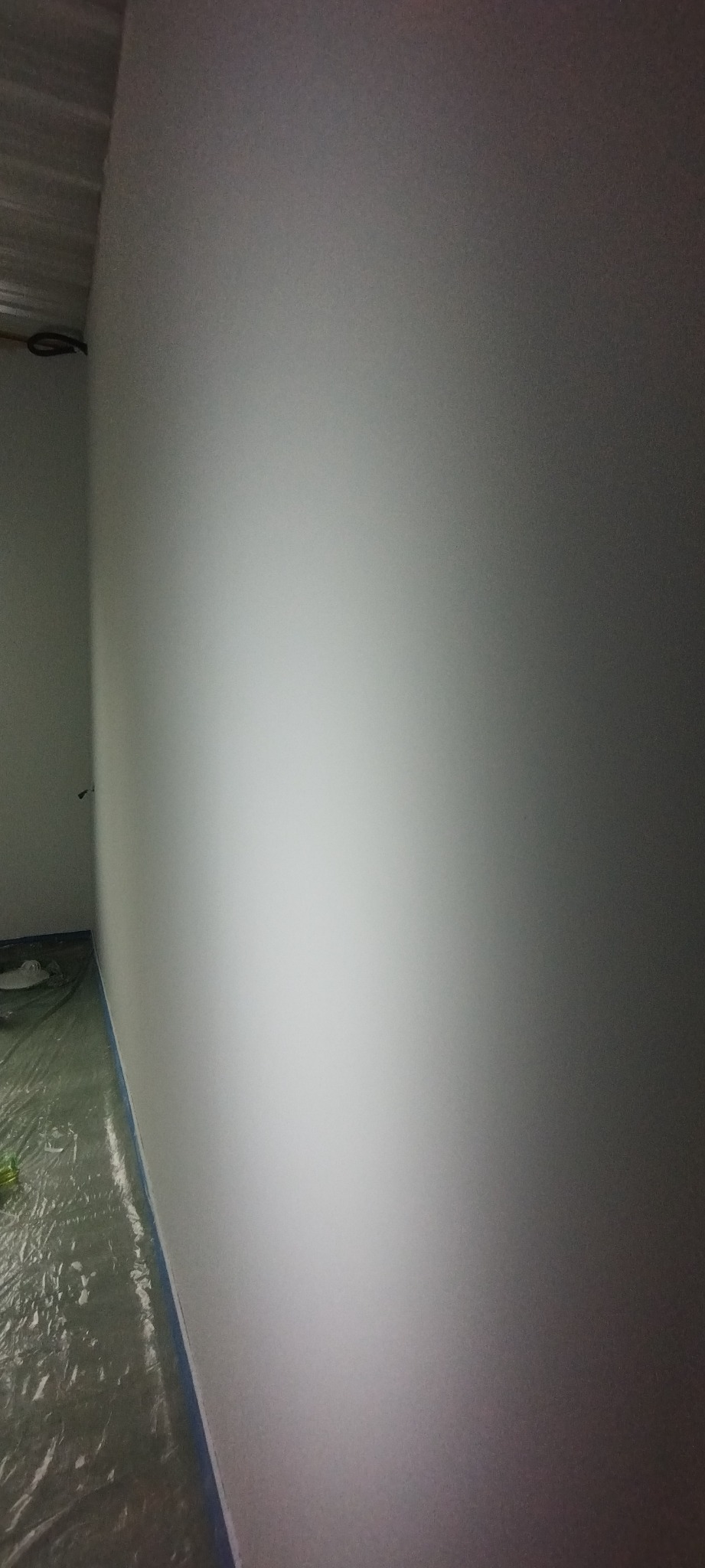 Świeżo pomalowana na biało ściana, zabezpieczona folią malarską na podłodze, widoczne ślady taśmy malarskiej przy podłodze.
